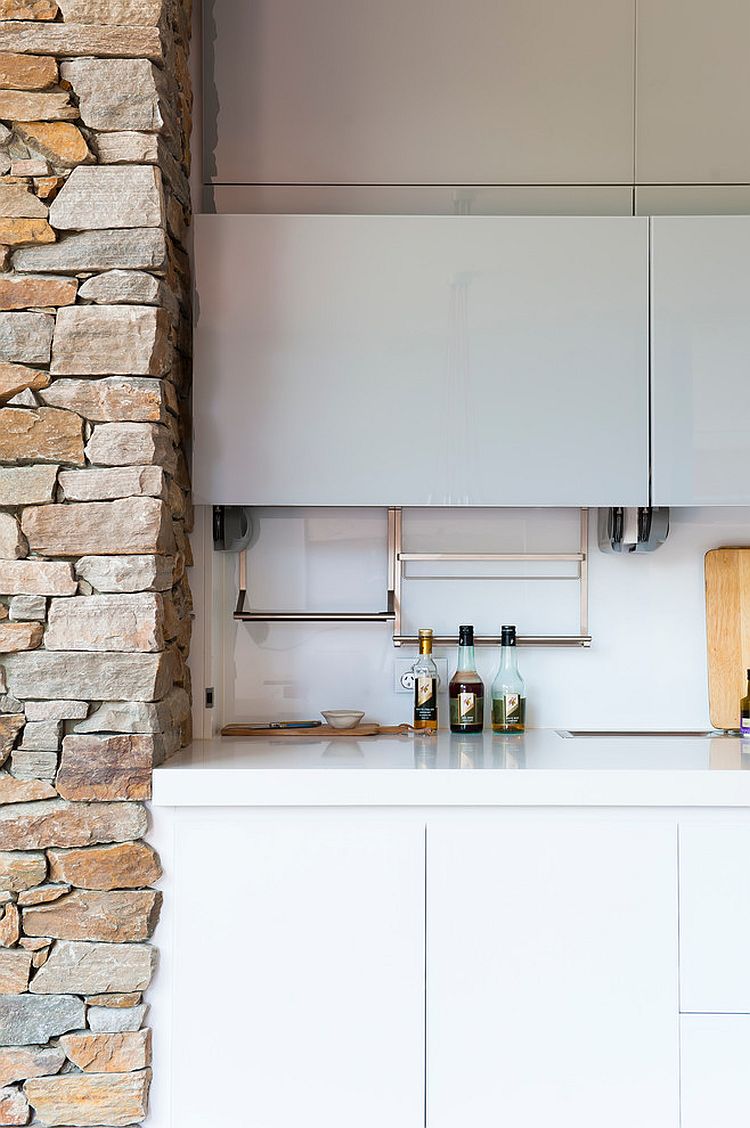 Stone fireplace interacts with glass white shelves of the kitchen