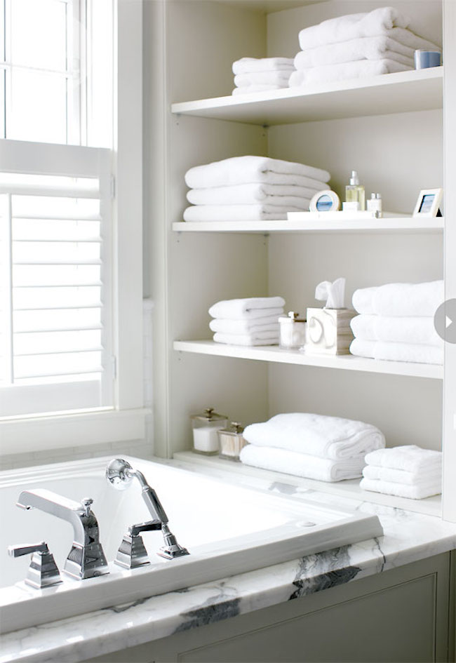 Open shelving at end of bathtub in white, chic bathroom