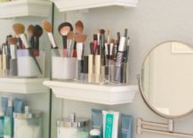 Three small white floating shelves in a bathroom that carry makeup brushes and skin care products.