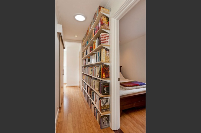 Hallway with tall bookshelves filled with books and a room with a bed.