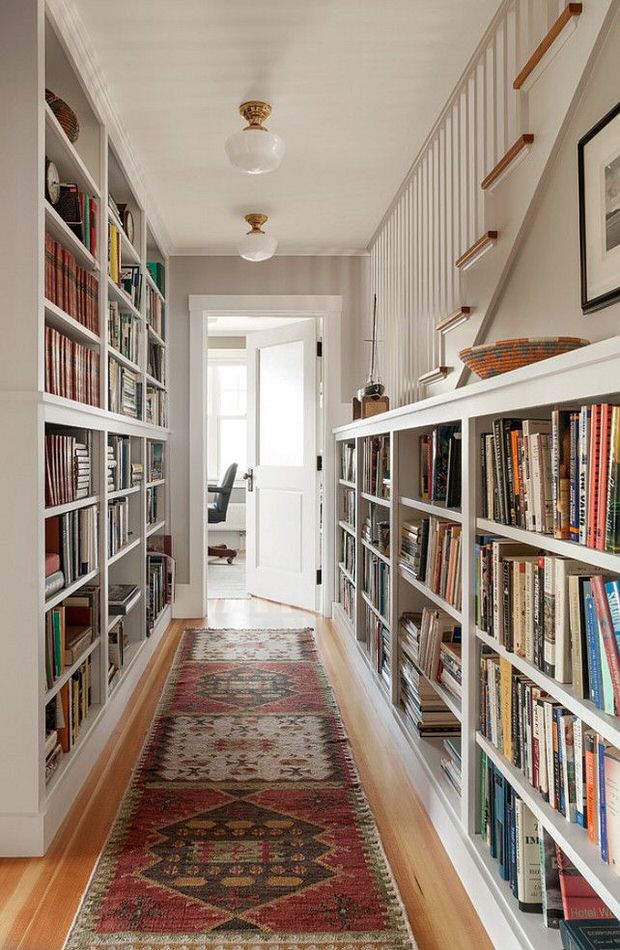 A long hallway stretches ahead, adorned with tall bookshelves filled with books on both sides split by a rug that lies on the floor.