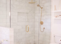 A glass and brass shower enclosure is filled with marble tiles lined with a Kohler Rainhead with Katalyst Air-Induction Spray in Vibrant Moderne Brushed Gold and a Purist Multifunction 3-Way Handshower.
