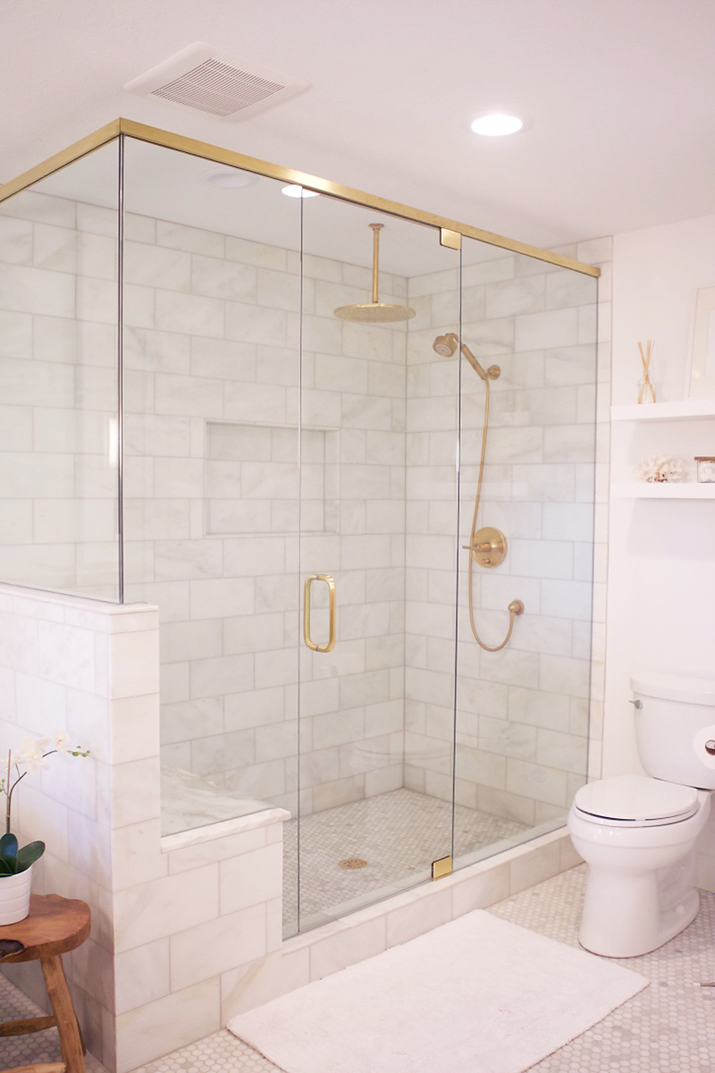 A glass and brass shower enclosure is filled with marble tiles lined with a Kohler Rainhead with Katalyst Air-Induction Spray in Vibrant Moderne Brushed Gold and a Purist Multifunction 3-Way Handshower.