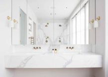 Modern bathroom features a floating marble vanity fitted with his and her sinks and gold faucets under mirrored medicine cabinets illuminated by white glass and gold sconces.