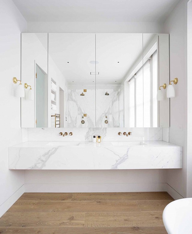 Modern bathroom features a floating marble vanity fitted with his and her sinks and gold faucets under mirrored medicine cabinets illuminated by white glass and gold sconces.