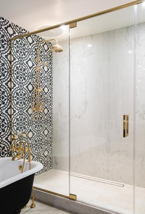 White and black bathroom features an accent wall clad in black and white cement tiles, Cement Tile Shop Bristol Tiles, lined with a black clawfoot tub fitted with a shiny brass vintage hand held tub filler illuminated by a Suzanne Kasler Morris Lantern placed next to a glass and brass shower enclosure.