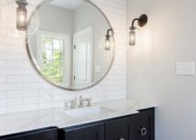Black and white bathroom boasts a black washstand complemented with large nickel ring pulls and a marble countertop fitted with a sink accented with polished nickel faucet located under a large round vanity mirror mounted on a white linear tile backsplash and lit by facing bronze and glass globe wall sconces.