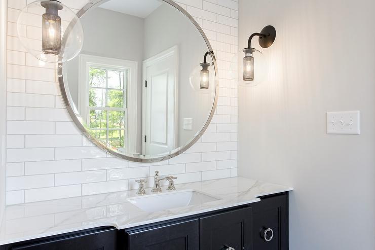 Black and white bathroom boasts a black washstand complemented with large nickel ring pulls and a marble countertop fitted with a sink accented with polished nickel faucet located under a large round vanity mirror mounted on a white linear tile backsplash and lit by facing bronze and glass globe wall sconces.