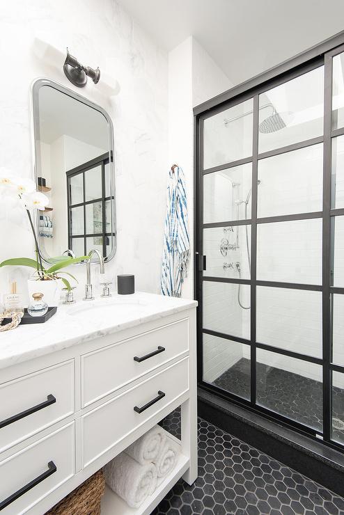 Restoration Hardware Astoria Flat Mirrors are lit by a white glass and bronze 2 light wall sconces mounted above a white dual washstand accented with a towel shelf, bronze pulls, and a honed white marble countertop fitted with a sinks and polished nickel gooseneck faucets.