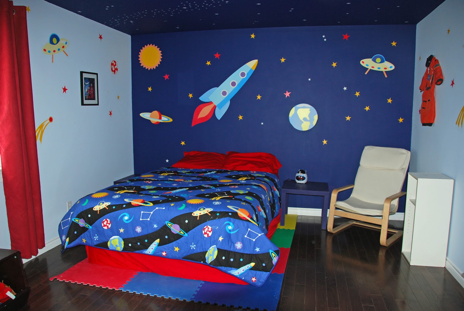 Cama colocada em quadrados de espuma ladeados por arte de parede espacial, incluindo um foguete, OVNI, Terra e sol.