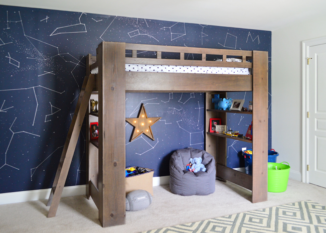 Quarto infantil com tema espacial, constelações pintadas na parede e uma cama elevada que funciona como área de recreação embaixo.