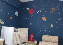 Space nursery with a cartoon-like space theme and a white crib beside a chair with a low back.