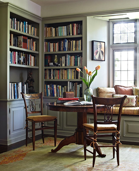A cozy room with a wooden table and two chairs placed in front of a large window. The table is adorned with a vase of fresh flowers and books.