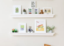 Two rolling wooden boxes flanked by three white floating shelves that hold picture frames, planters, and toys.