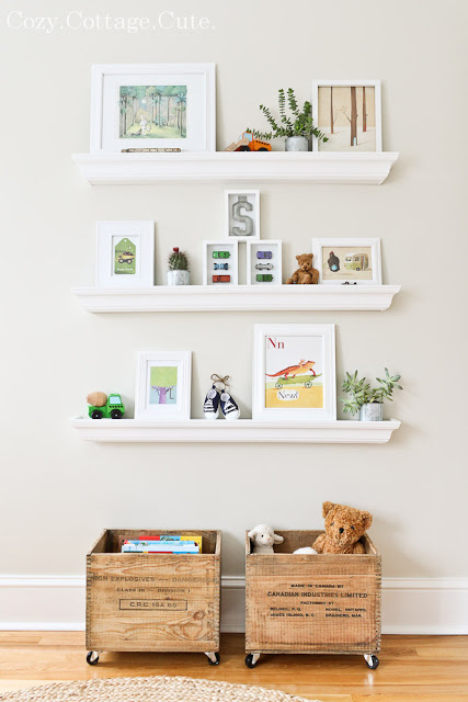 Two rolling wooden boxes flanked by three white floating shelves that hold picture frames, planters, and toys.
