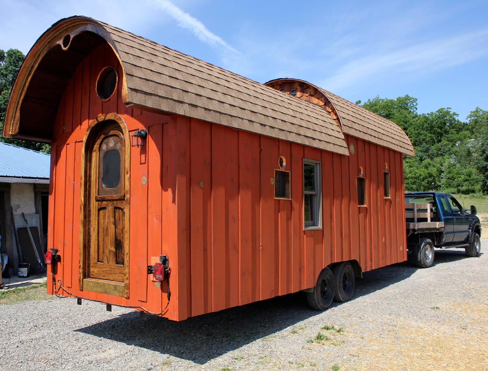 Red exterior of a trailer-style tiny home, featuring a roof made of shingles.