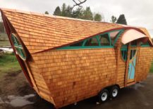 Uniquely shaped tiny home on a trailer frame, which has a roof made of shingles that spans almost the entire way down and has green accents.
