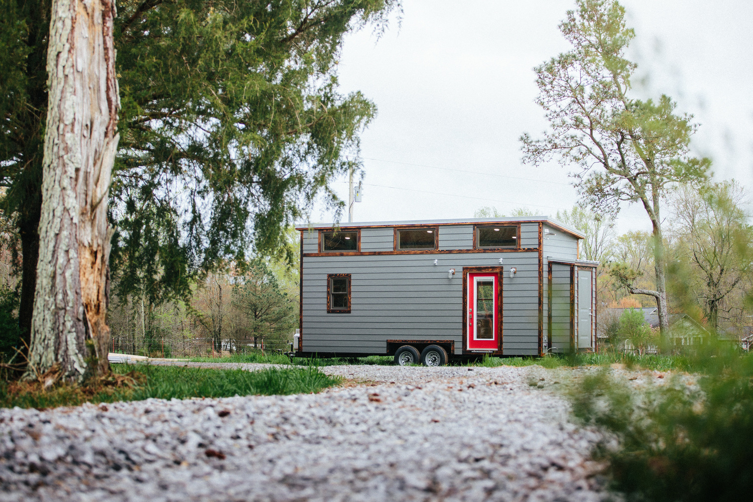 Sleek tiny home on a trailer frame, showcasing grey color with wood accents that clash with the red front door.