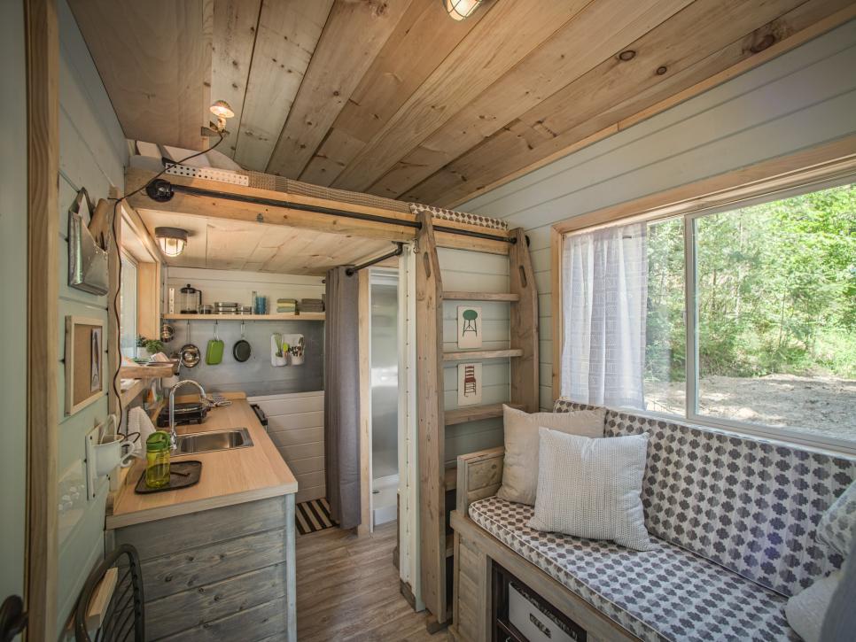 Modern interior of a tiny home which has wood finishes all over, kitchen and seating on the first level and a bed with lamp on the second level.
