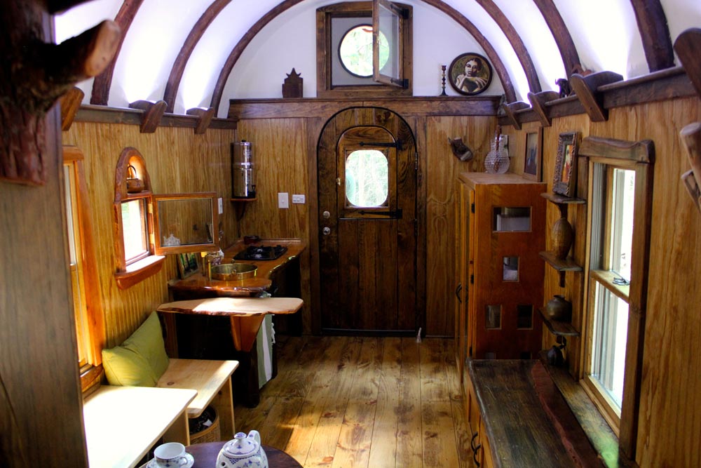 Interior of tiny home with wood floors and walls, white ceiling with wood beams, and a unique wood front door.