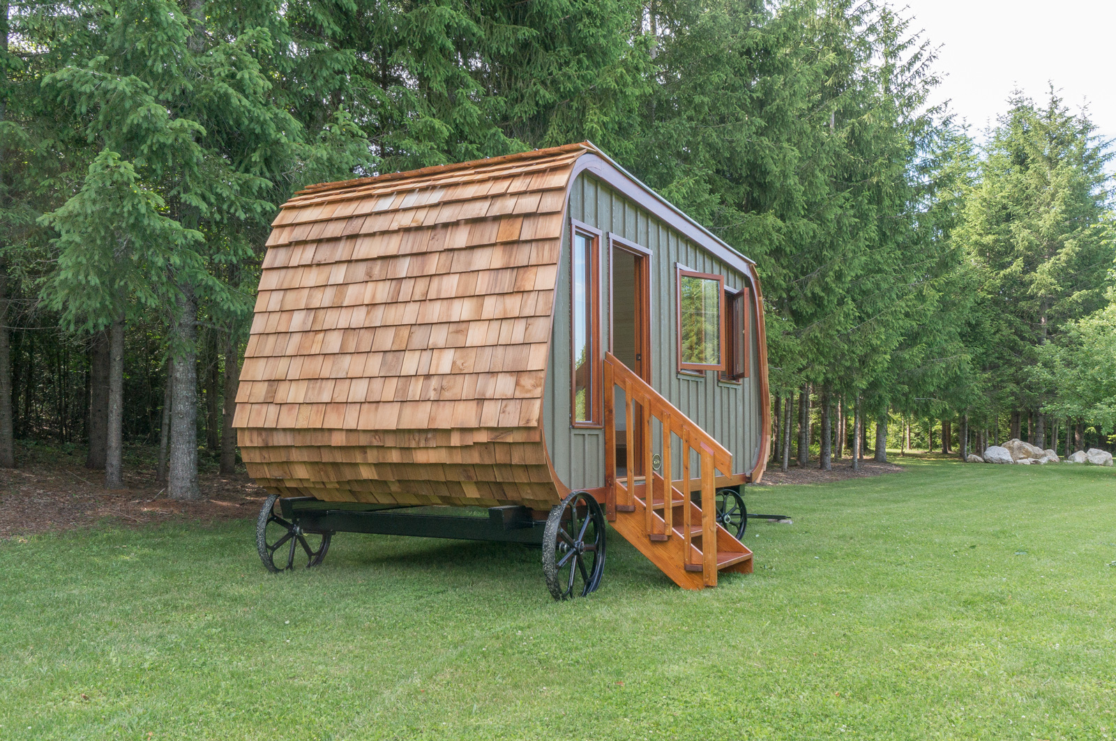 Tiny home on a trailer frame with a cart look on large metal wheels, featuring a roof that wraps around and green sides with large windows that has wood frames.