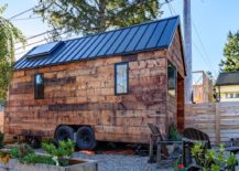 Trailer-style tiny home with wood board exterior, tiny windows, metal roof with skylight, and a small garden.