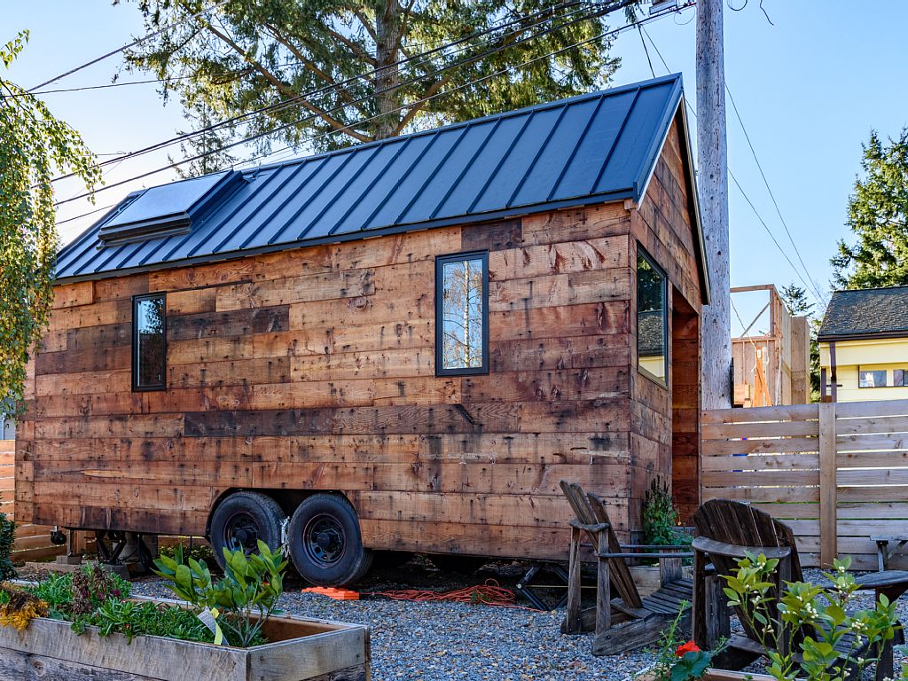 Trailer-style tiny home with wood board exterior, tiny windows, metal roof with skylight, and a small garden.