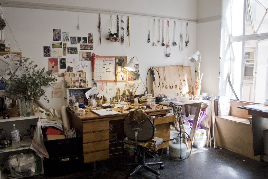 A desk with tons of art supplies in an art studio with hanging art on the wall.