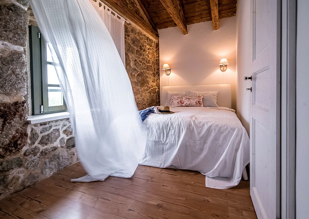 Stone wall and wooden ceiling coupled with white sheer curtains add Mediterranean beauty to the small bedroom