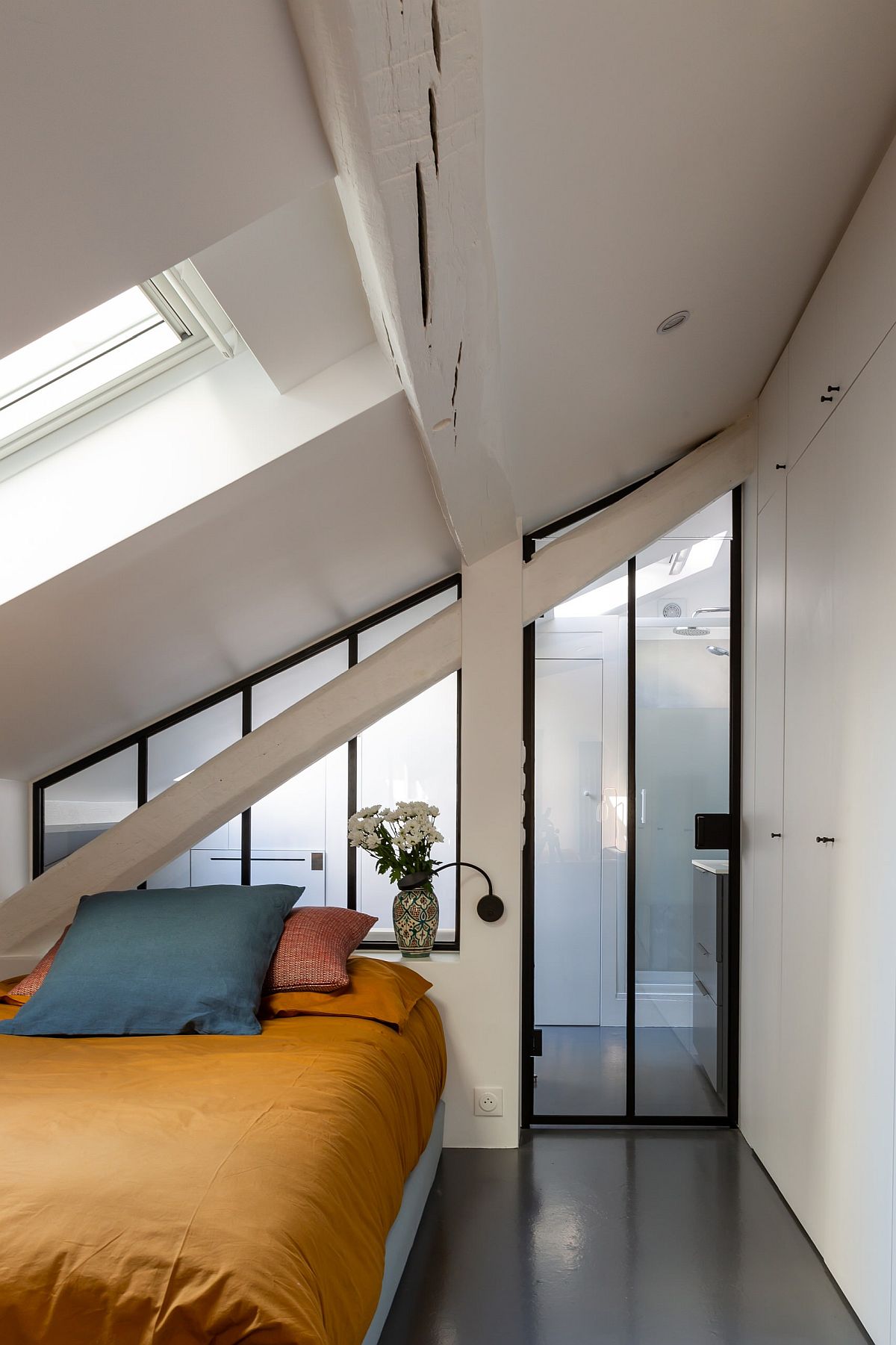 Light-filled small bedroom of French apartment with white walls, skylights, gray floor and colorful bedding