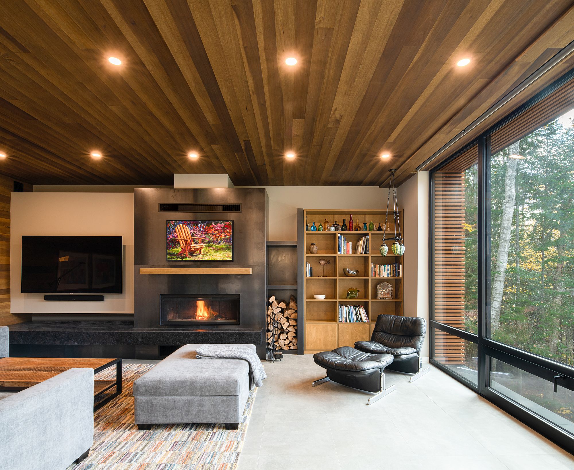 Living area of the house with a smart fireplace, woodsy ceiling and large floor-to-ceiling glass walls that bring the outdoors inside