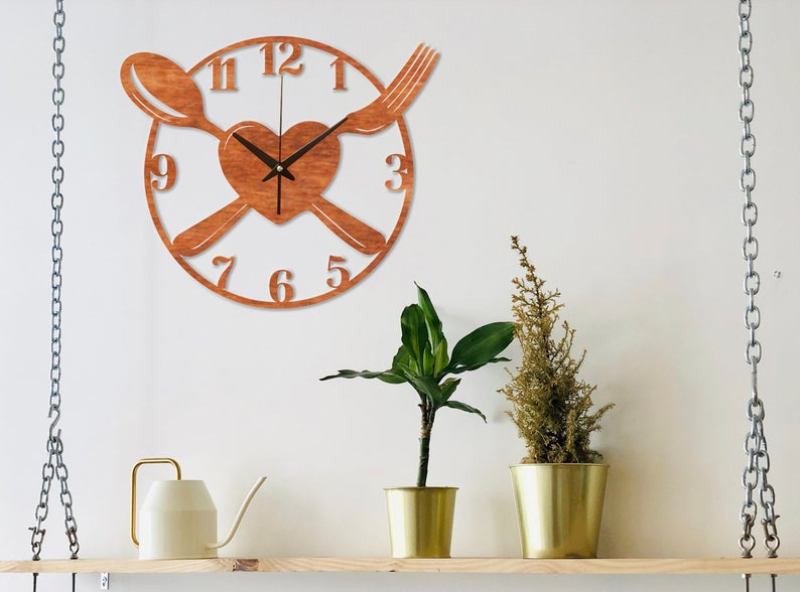 Kitchen wall clock with spoon, fork and heart design hanging on top of a shelf with plants