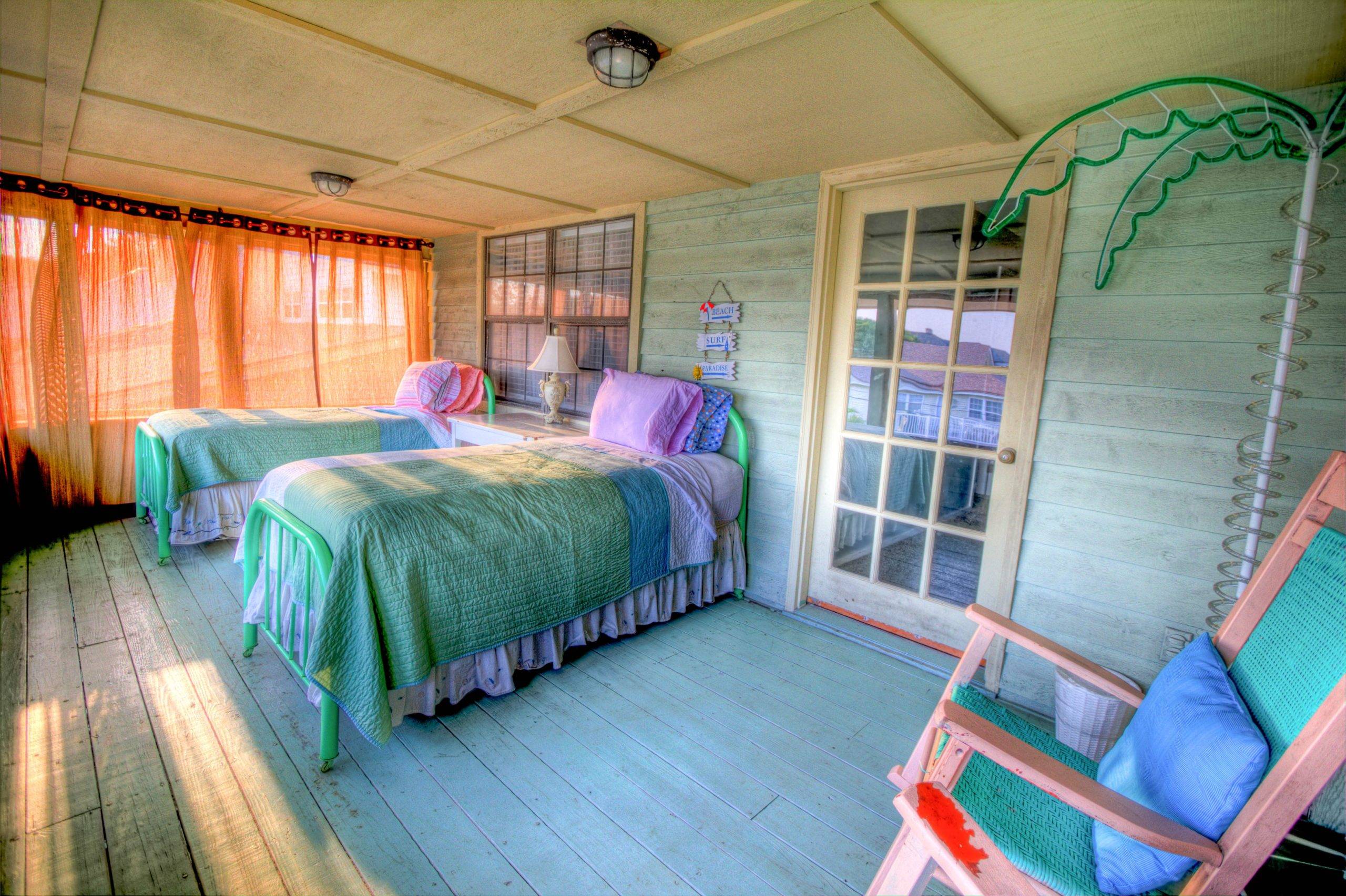 Two beds in spacious bedroom with orange sheer curtain and palm tree stand at the side