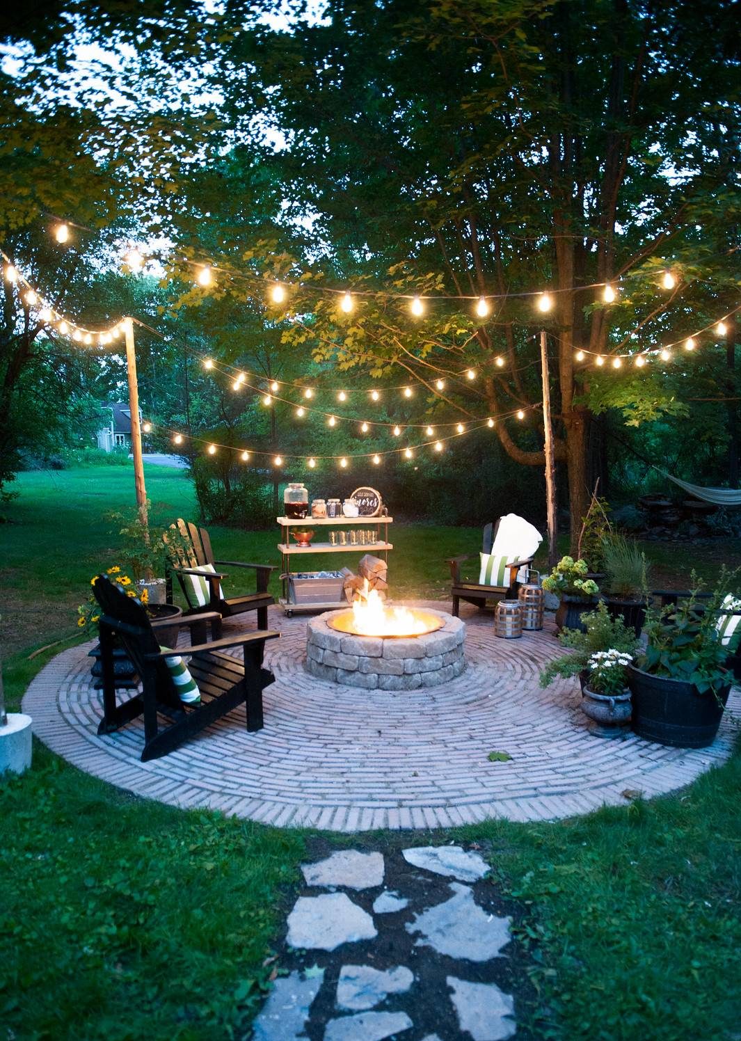 Outdoor fire put surrounded by black chairs, plants and dangling lights