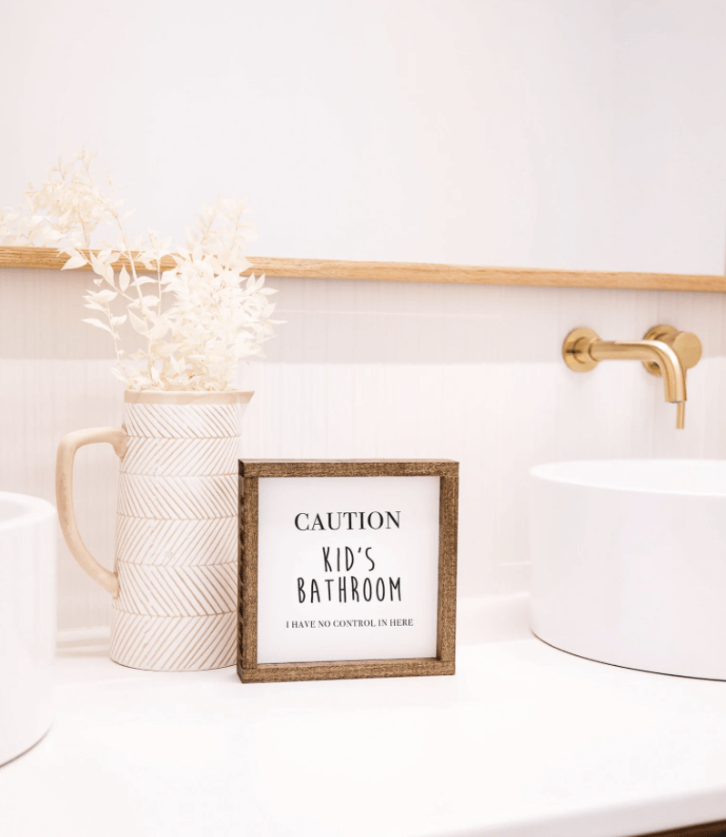 caution kids bathroom rustic wood frame sign with white background on white counter gold faucet pitcher with stems