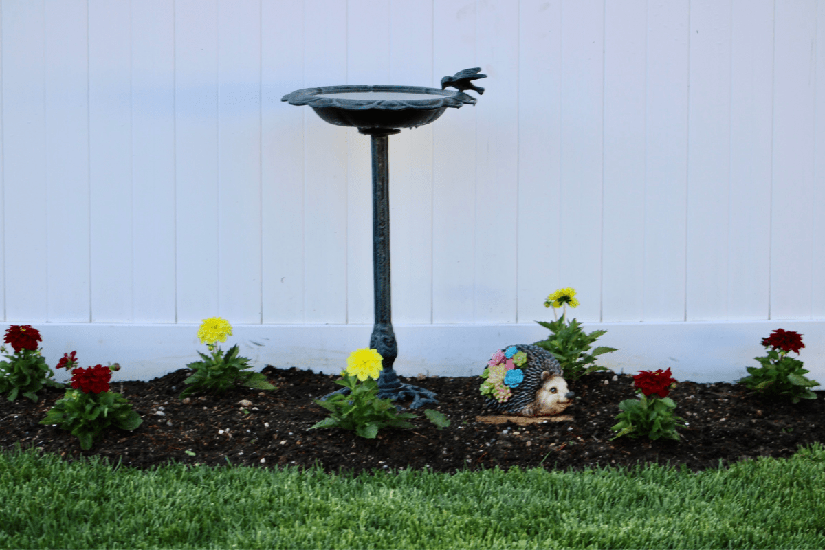 bird bath in front of white fence with flowers in garden grass