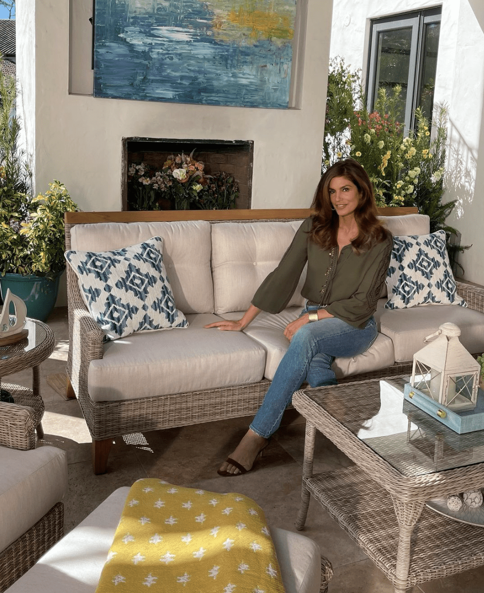 cindy crawford sitting on a outdoor couch with pillows in front of fireplace