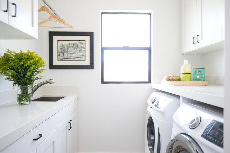 white laundry room side by side washer dryer golden rod jar sink black frame window clothes drying rod white shaker cabinets