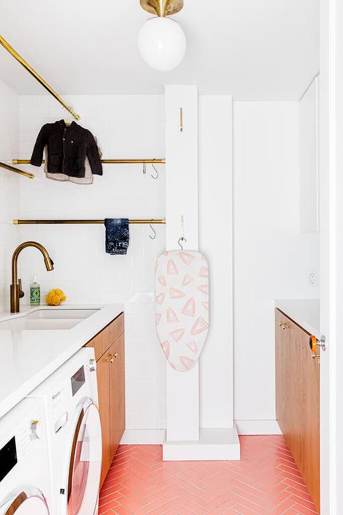 modern laundry room pink herringbone floor tiles brown flat front wood cabinets brass hardware sink with antique brass faucet drying rods white subway tile front loading washer and dryer glass globe semi flush mount