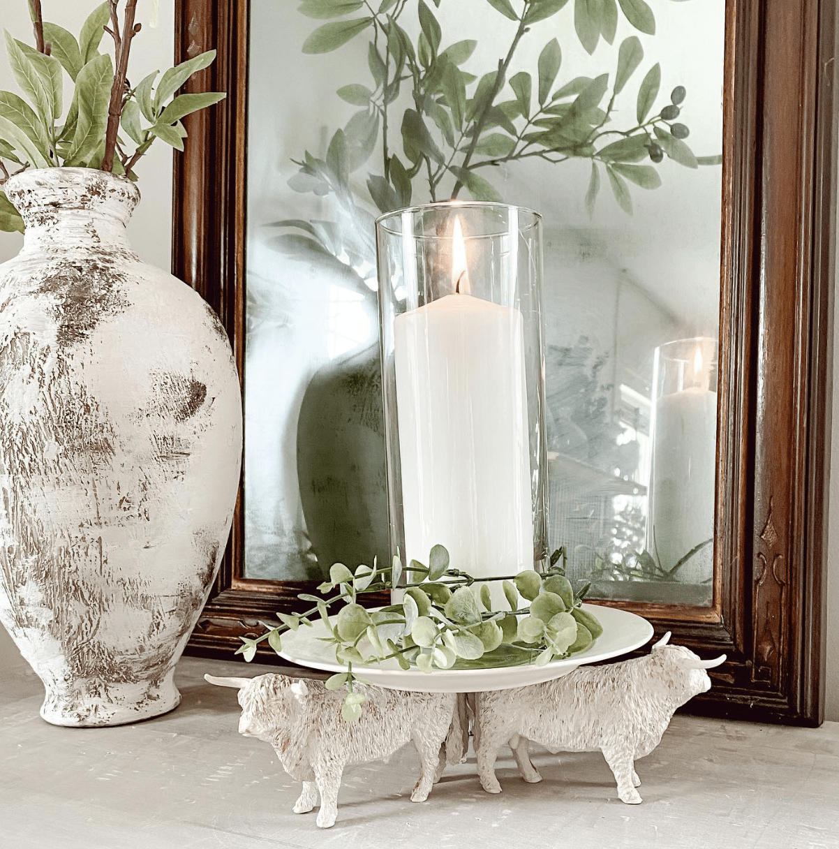 white candle holder with highland bulls pillar candles on table with mirror behind and rustic vase