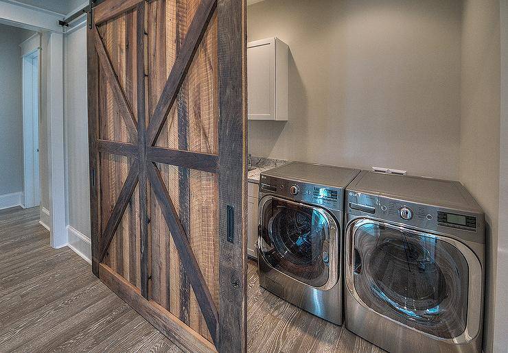 side by side silver washer and dryer behind rustic barn wood sliding door grey walls