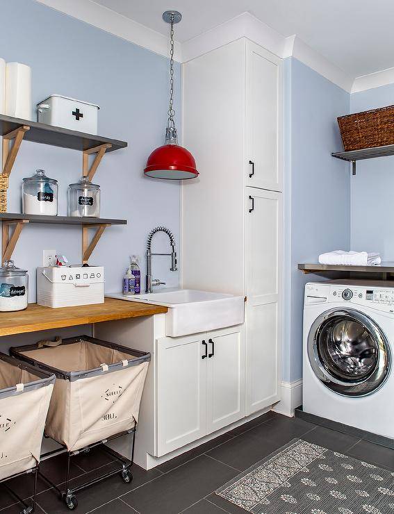 blue wall small laundry room with black tile floor wall mounted gray shelves on wood brackets red antique lantern white shaker cabinets roll out laundry hampers white washer