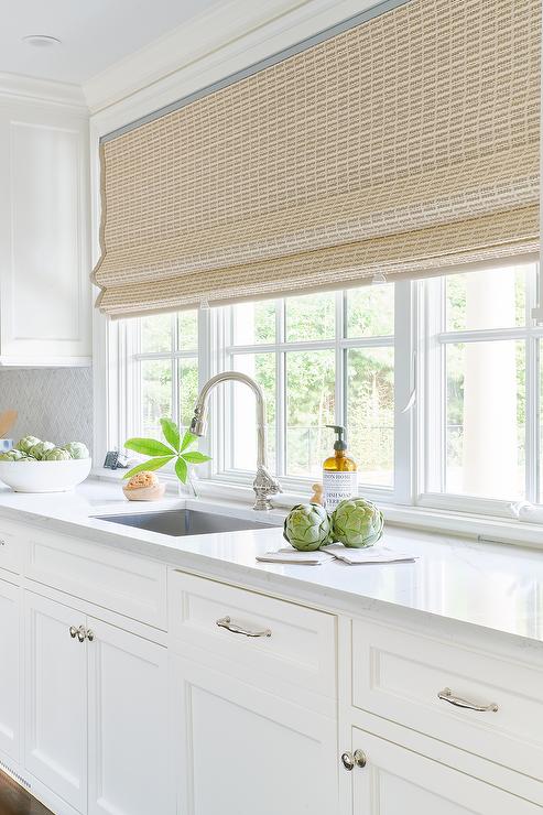 A row of windows are covered in a tan bamboo roman shade hung over a sink with a polished nickel gooseneck faucet mounted to a light gray quartz countertop accenting white kitchen cabinets