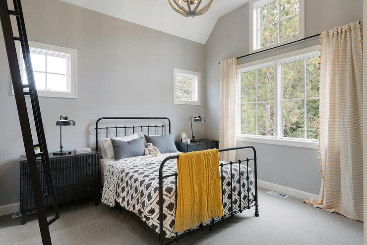 lack and gray boy's bedroom black dressers topped with oil rubbed bronze task lamps and placed beneath windows on either side of an iron bed. The bed is dressed in black and white bedding and sits on gray carpeting in front of a gray wall. The window beside the bed is covered with yellow striped pom pom curtains.