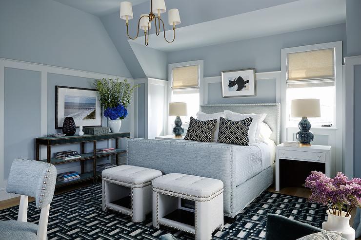 gray and blue bedroom boasts white nailhead stools placed on a blue and black geometric rug at the foot of a heather gray bed complemented with black geometric pillows. Art hangs over the bed from a blue wallpapered wall finished with white moldings. Blue double gourd lamps sit atop a white waterfall nightstand located beneath windows.