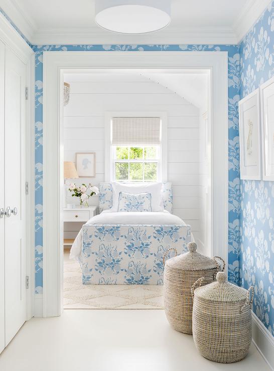 White and blue cottage style bedroom features a white and blue floral print twin bed placed against a white shiplap wall and under a window covered in a bamboo roman shade. A white farmhouse nightstand is adorned with brass knobs, while the bed sits on a cream Moroccan trellis rug.