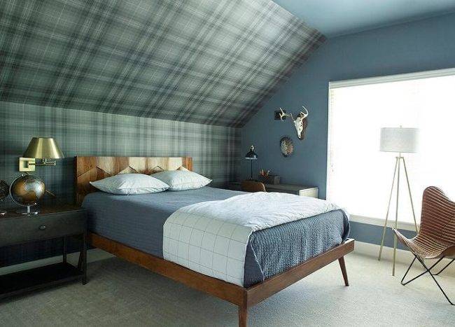 A two-tone nightstand lit by a brass swing arm sconce is placed beside a reclaimed wood bed and placed against a wall clad in green and brown plaid wallpaper leadup to a wallpapered sloped attic ceiling. Perimeter walls are covered in a gorgeous blue paint.