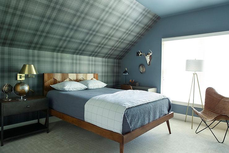 A two-tone nightstand lit by a brass swing arm sconce is placed beside a reclaimed wood bed and placed against a wall clad in green and brown plaid wallpaper leadup to a wallpapered sloped attic ceiling. Perimeter walls are covered in a gorgeous blue paint.