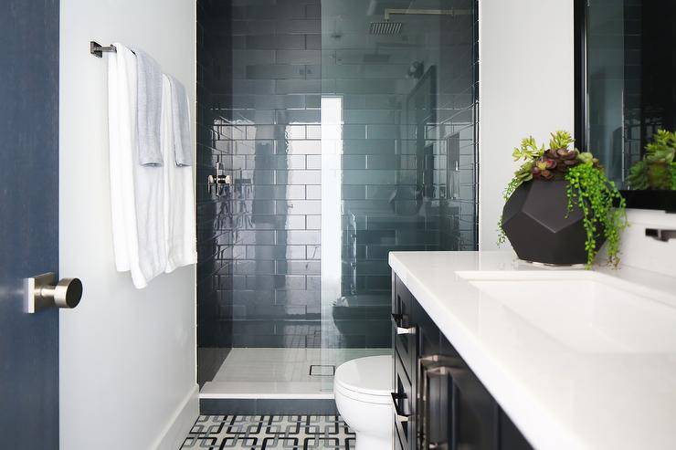 Black and blue geometric floor tiles black washstand with a white quartz countertop vanity mirror nickel towel bar mounted to a wall beside a walk-in shower finished with a glass partition and glossy blue stacked wall tiles