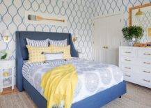 yellow and blue bedroom features an oar art piece hung from a wall covered in blue trellis wallpaper and mounted above a blue wingback bed. The bed, flanked by brass sconces mounted over white oval nightstands, is dressed in blue medallion bedding accented with yellow pillows